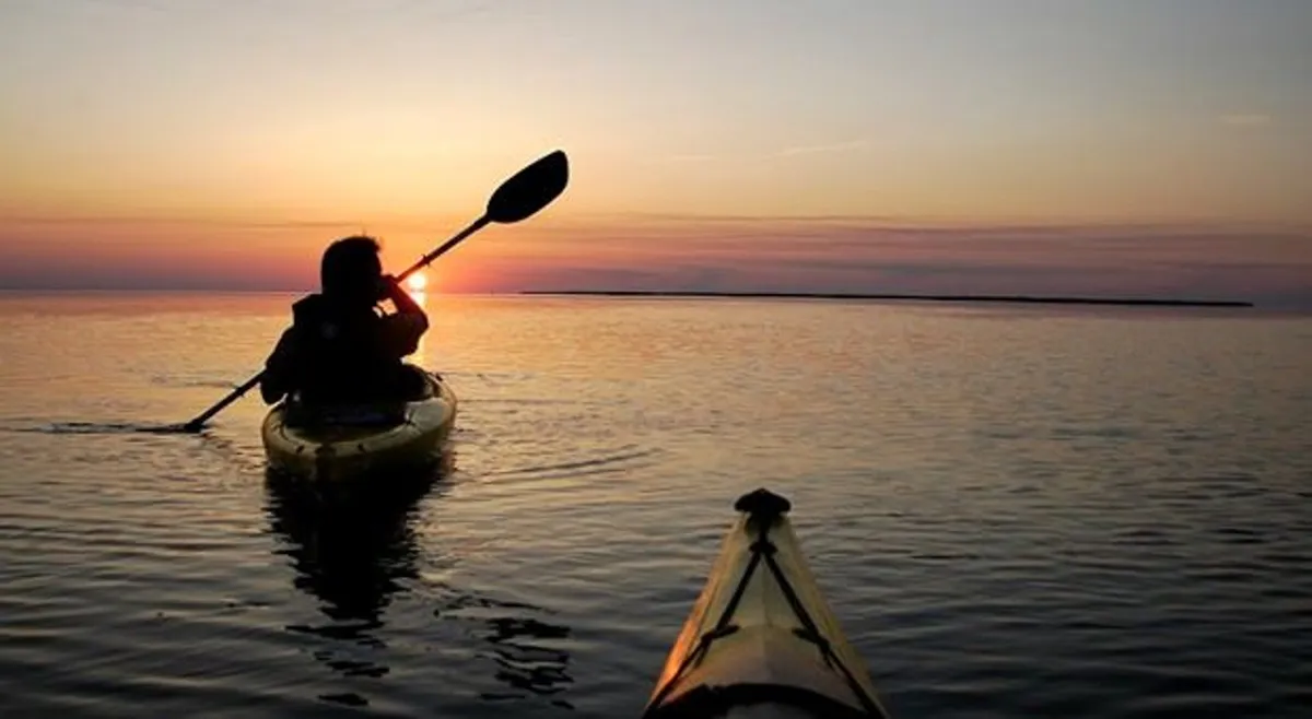 A tranquil scene of a kayaker silhouetted against the setting sun, gliding across calm waters. The peaceful mood and natural beauty evoke the spirit of exploration on Currituck Sound.