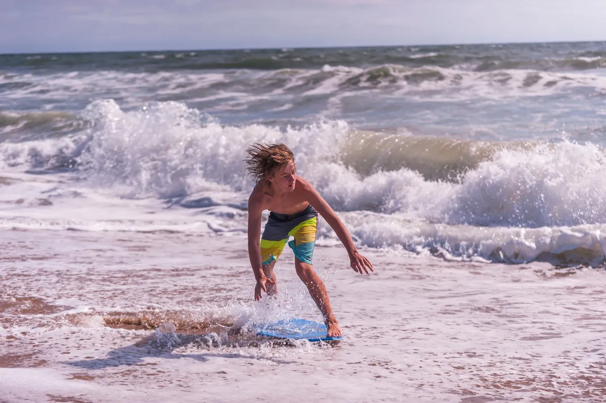 A joyful beach scene with a boy riding a boogie board on the waves, soft sand underfoot, and laughter shared among friends in the background. The sun is setting, casting a warm glow over the shoreline, and the scene hints at group activities like swimming, beach games, and even the promise of bonfires at dusk.