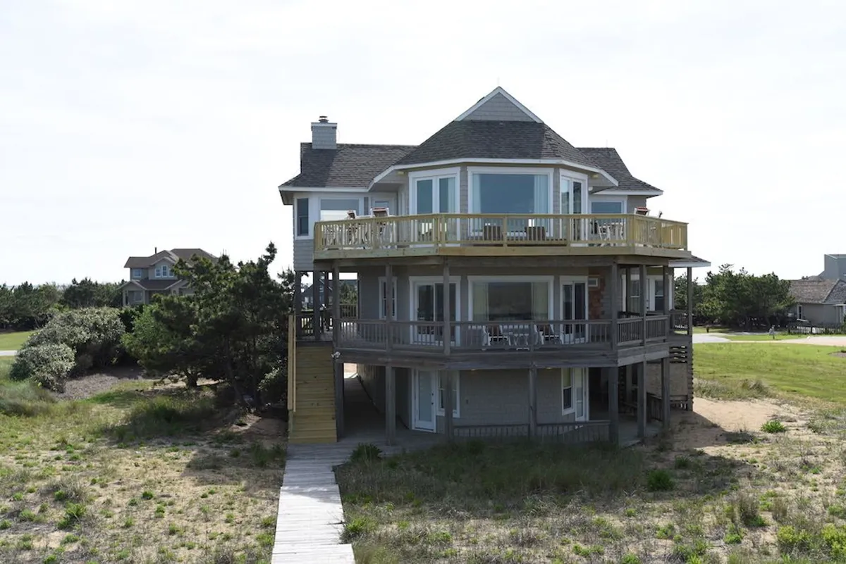 A wide, sunlit deck stretches along the home’s exterior, bordered by lush greenery. Expansive windows invite the outdoors in, while groups of people enjoy relaxed outdoor dining and casual gatherings. The setting is perfect for sunrise coffees, sunset barbecues, and late-night stargazing.