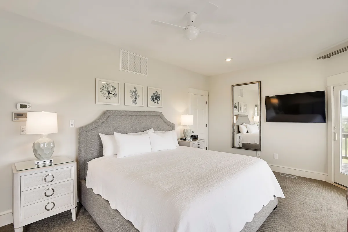 A bright and tranquil bedroom featuring a plush bed adorned with soft linens, flanked by stylish side lamps. The room includes a large mirror, a modern television, and tasteful wall art, with natural light streaming in from a window.