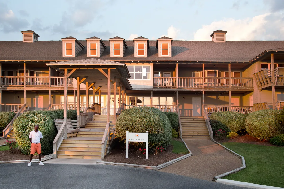 A scenic view featuring a staircase leading to the entrance, surrounded by large round bushes and a signpost that reads 'A Beach House.' People can be seen relaxing on the porch, socializing with friends, and enjoying sunset views, conveying a welcoming, tranquil atmosphere.
