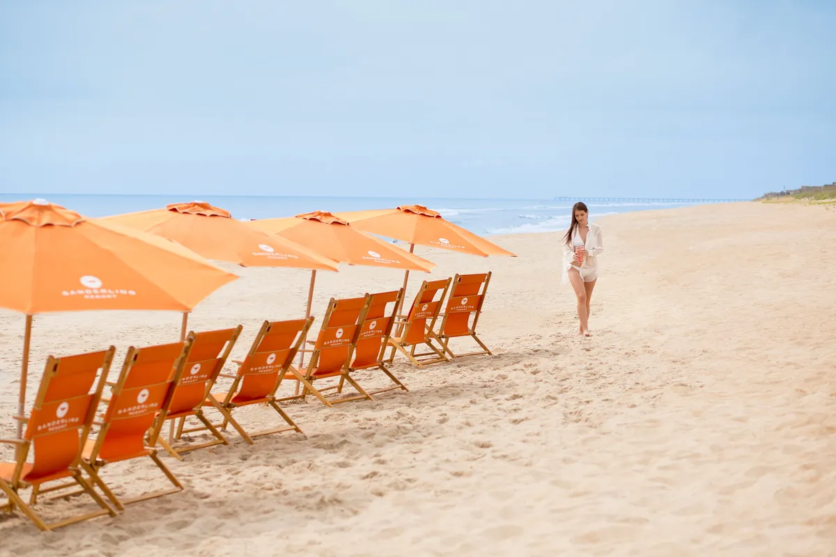 A wide view of a sandy beach with colorful beach chairs and umbrellas scattered along the shore. The ocean stretches out beyond, and a woman strolls near the water, evoking a peaceful, family-friendly beach atmosphere.