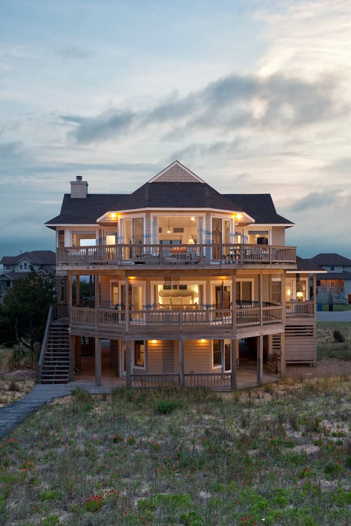 A welcoming exterior shot of a multi-level coastal home. The image captures spacious decks with outdoor seating, large windows that invite natural light, and a beautifully landscaped yard. The setting sun casts a warm glow, inviting thoughts of evening relaxation and outdoor dining.