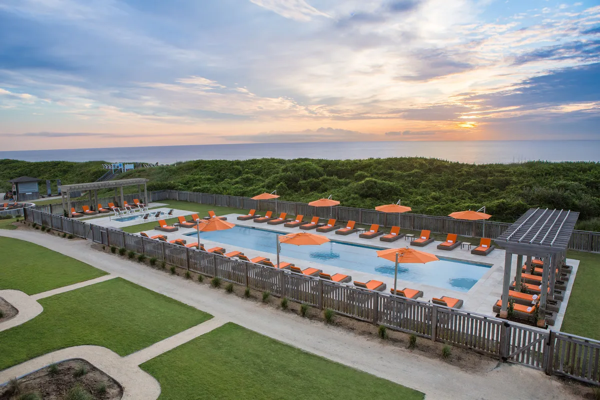 A sparkling swimming pool surrounded by orange umbrellas and lounge chairs, with the sun setting over the ocean in the background. Guests are seen swimming and relaxing, capturing the joyful spirit of a day by the water.