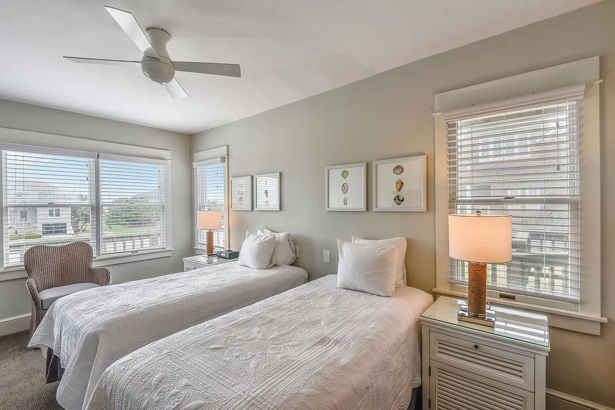 A serene bedroom with twin beds, each topped with crisp linens. The room is softly lit by a bedside lamp and natural light from the windows, featuring an armchair and wall artwork for added comfort.