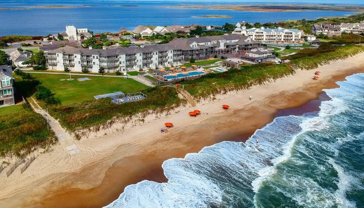 An inviting beachfront scene shows a swimming pool surrounded by comfortable loungers, with a hotel complex and rows of beach umbrellas just steps from the shore. Guests are seen sunbathing, swimming, and casually gathering, all set against the tranquil backdrop of the ocean.