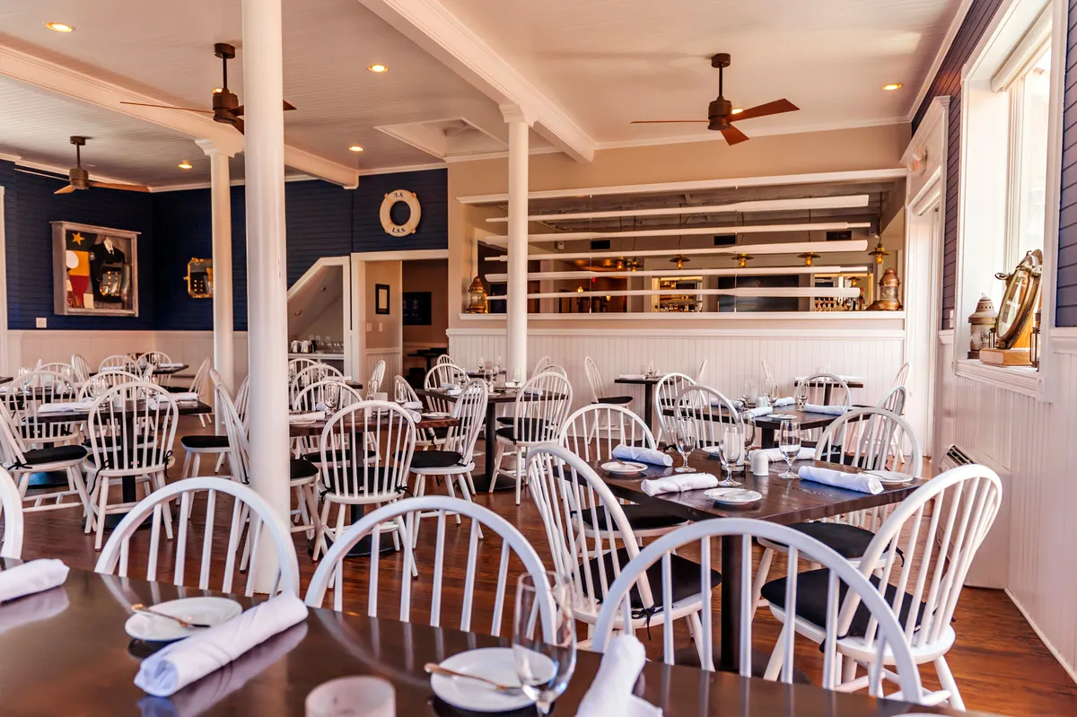A bright interior featuring white wooden chairs paired with dark wooden tables. Decorative wall art and a visible bar area create a friendly, welcoming environment. The arrangement offers ample seating for group meals and social gatherings.