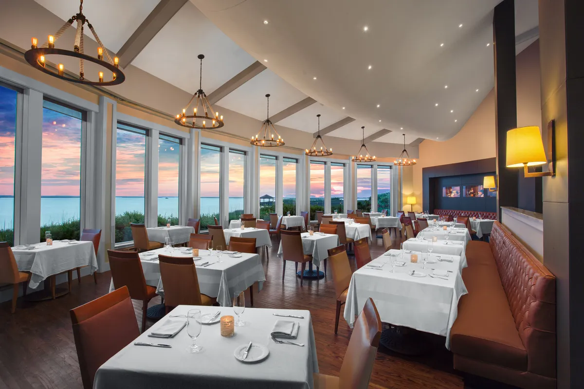 A dining area with large windows capturing a stunning sunset view. The tables are elegantly set with fine dinnerware, and chandeliers provide a warm, ambient glow. The space feels inviting and sophisticated, ideal for gatherings and celebratory meals.