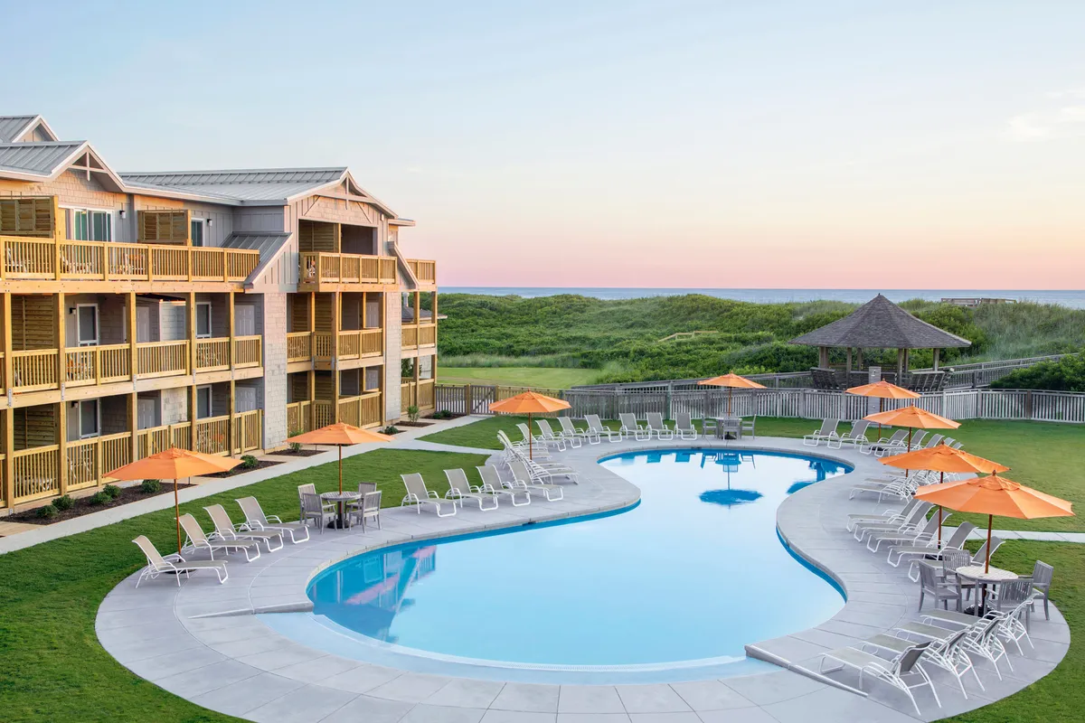 A sun-drenched swimming pool area featuring lounge chairs and bright orange umbrellas, set against the backdrop of the building facade. People are swimming, sunbathing, and socializing, highlighting both leisure and resort amenities.