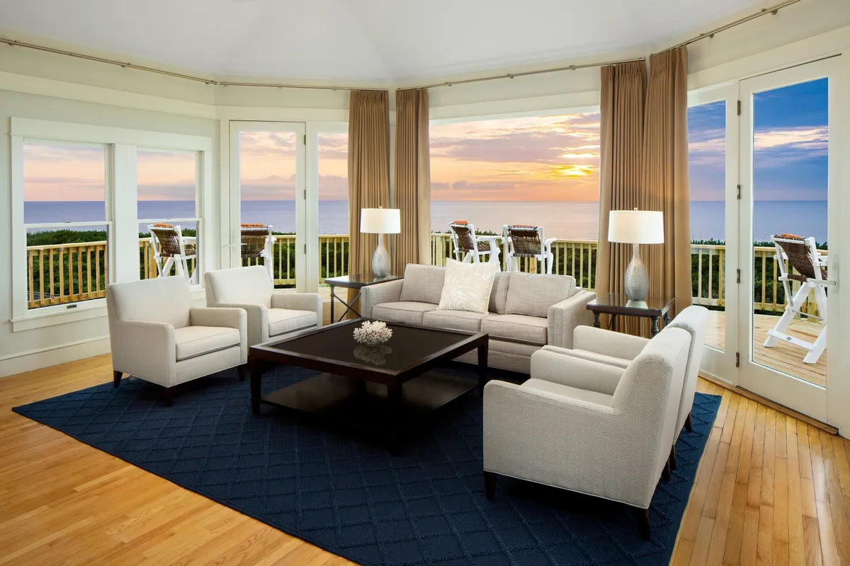 The image showcases a cozy and inviting living room with large windows offering a breathtaking sunset view over the Currituck Sound. Plush sofas and armchairs are arranged around a stylish coffee table with a decorative centerpiece, creating a perfect space for relaxation or gathering with loved ones.