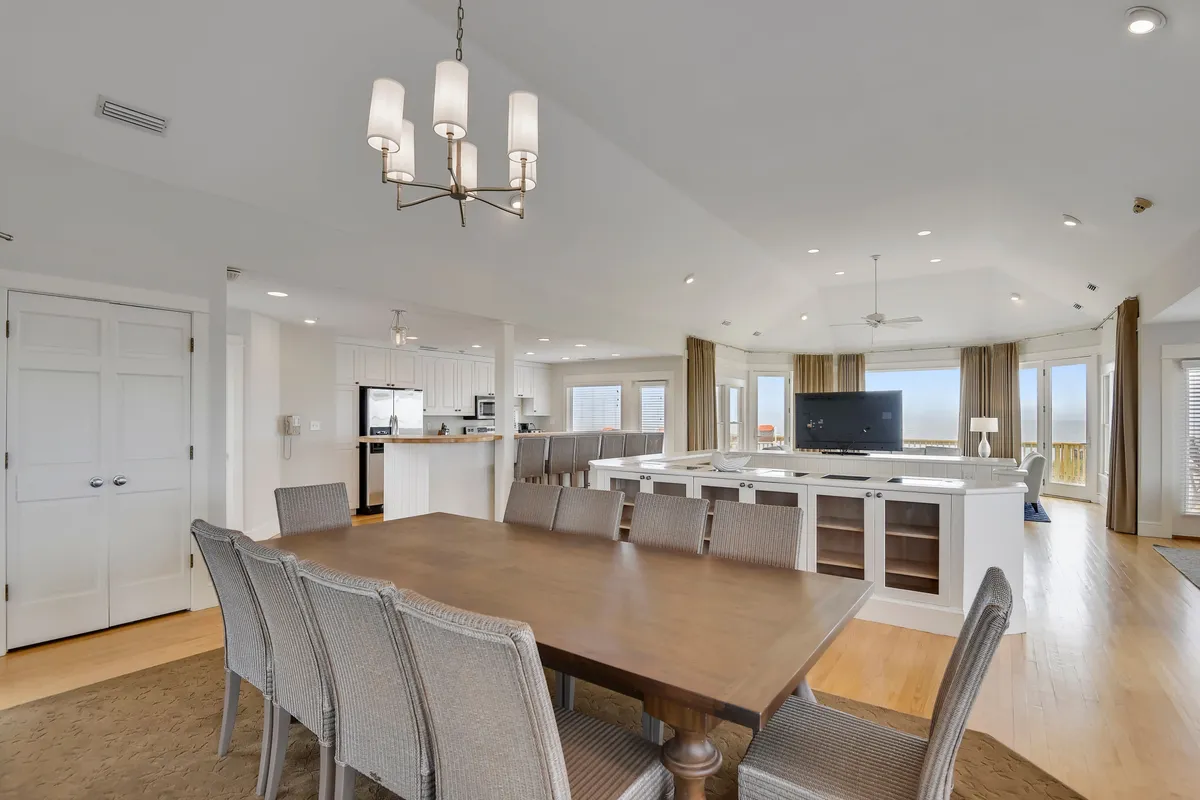 A modern, airy space blending a dining area and part of a living area. The room is anchored by a large dining table, flanked by plush chairs and surrounded by expansive windows. A modern kitchen and elegant chandelier complete the scene, creating a seamless flow between entertaining and relaxing.